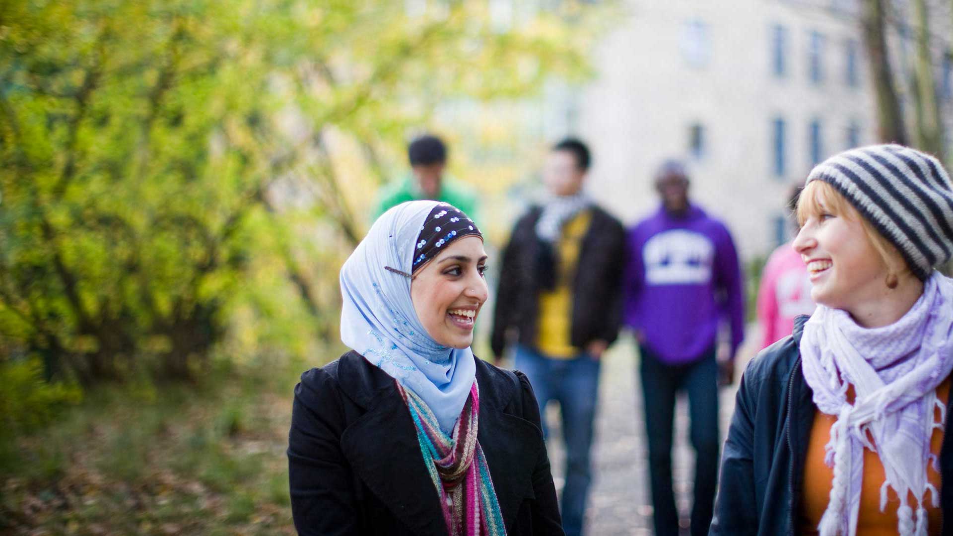Two students walking together outside, smiling and chatting. Another group of students are blurred in the background.